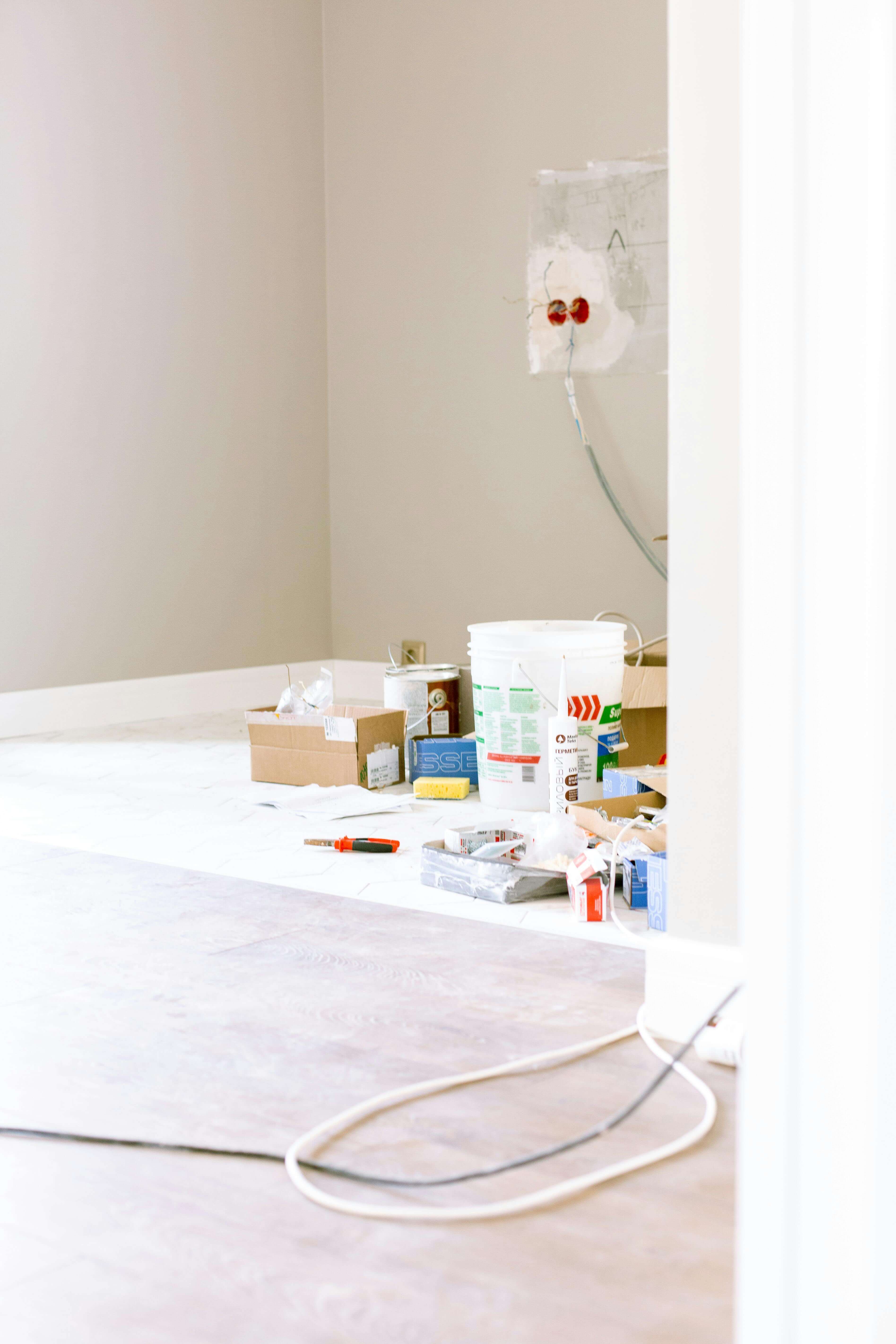 Huone remontin keskellä, jossa maalaustarvikkeita, sähköjohtoja ja lattialaminaattia. Valoisa ja avara tila.